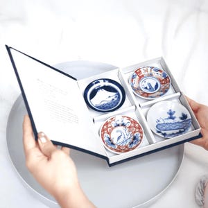 Può includere: Un set di quattro piccoli piatti rotondi in ceramica con disegni blu e rossi, presentati in una scatola bianca con bordo blu navy. Ogni piatto misura circa 10 cm di diametro.