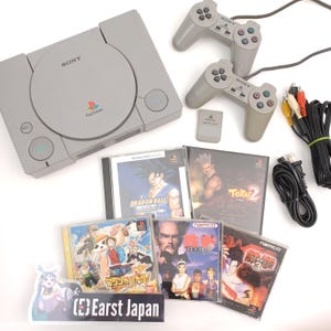May include: A gray Sony PlayStation console with two matching controllers, a memory card, and several game cases. The game cases feature colorful graphics and titles like Dragon Ball and Tekken. Cables and power cords are also included.