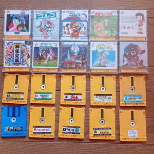 May include: A collection of vintage Nintendo Famicom game cartridges and floppy disks. The game cartridges feature colorful artwork and titles in Japanese. The floppy disks are yellow and blue, with labels.