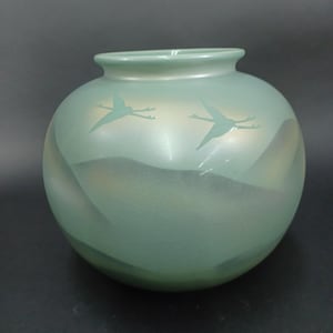 May include: A round, seafoam green vase with a wide opening. The vase features a design of two birds in flight, etched into the surface. The background has a subtle, layered effect. The vase is a decorative item.