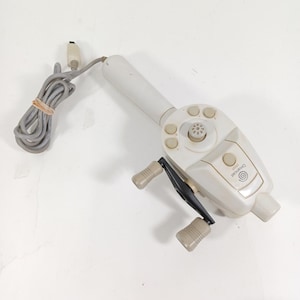 May include: A white Dreamcast fishing controller with a gray cable and a beige handle. The controller has several buttons and a reel with beige handles. The Dreamcast logo is visible on the controller.