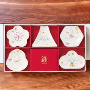 May include: A set of six white ceramic dishes with gold trim, each with a unique design, including floral and geometric patterns. The dishes are arranged in a red box with Japanese text. The box is inside a white container.