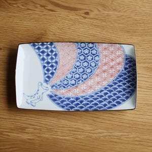 May include: Rectangular ceramic serving dish with a white base and a decorative pattern in blue and red. The design includes geometric shapes and a stylized animal figure. The dish has a dark brown border.