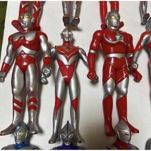 May include: A collection of Ultraman action figures featuring red and silver humanoid characters with distinct chest emblems, yellow or silver eyes, and smooth plastic bodies arranged in rows on a plain white surface for display.