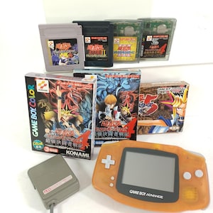 May include: A selection of retro Game Boy and Game Boy Advance items. Includes various game cartridges, boxes, and an orange Game Boy Advance console. The boxes feature colourful illustrations and Japanese text. A grey controller is also present.