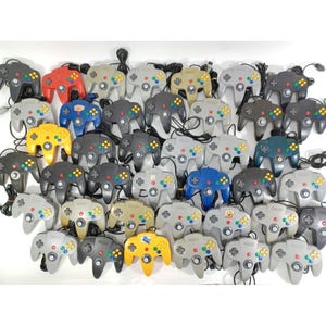 Könnte beinhalten: Eine Sammlung von Nintendo 64-Controllern in verschiedenen Farben, darunter Grau, Schwarz, Rot, Blau und Gelb. Jeder Controller hat ein unverwechselbares dreizackiges Design mit einem zentralen Joystick und mehreren Tasten. Die Controller sind mit schwarzen Kabeln verbunden.