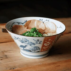 May include: A bowl of ramen with pork slices and green onions. The bowl has a white base with blue floral and bird patterns, and a red and white patterned section. The ramen is served on a wooden table.