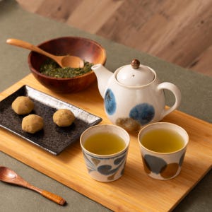 May include: A wooden tray with two blue and brown teacups filled with green tea, a small wooden bowl with green tea leaves, a wooden spoon, three brown mochi, and a white teapot with blue and brown spots.