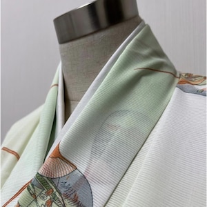 May include: Close-up of a kimono collar. The kimono features a white base with a light green and patterned design on the collar. The design includes orange and blue accents.