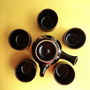 Puede incluir: Una tetera de cerámica negra y marrón con un diseño floral y cuatro tazas de té a juego. La tetera tiene un pico y un asa. Las tazas de té son redondas y no tienen asas.