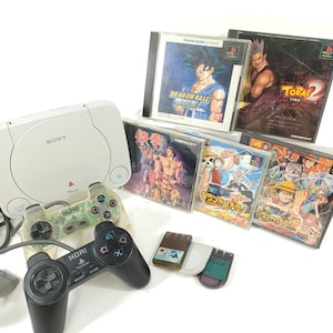 May include: A white Sony PlayStation console with two controllers and several game cases. The game cases feature colorful illustrations of anime characters and game titles. The controllers are black and clear.