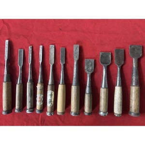 May include: A collection of eleven wood chisels with metal blades and wooden handles. The tools vary in width and length, featuring metallic ferrules at the base of the handles. Some handles display paper labels with Japanese characters.