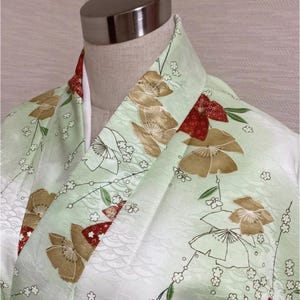 May include: A light green kimono with a floral pattern. The kimono features large, stylized flowers in shades of gold and white, with red accents. The fabric has a subtle wave pattern. The collar and trim are red.