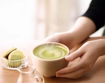 May include: A person holding a bowl of steaming green matcha tea. The bowl is made of light brown ceramic and has a rounded shape. The tea is a vibrant green color and has a swirl pattern on top.