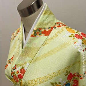 May include: A light green kimono with floral and geometric patterns. The kimono features red, yellow, and blue flowers, along with orange and gold accents. The garment is displayed on a mannequin, showcasing its design and fabric.