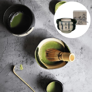 May include: An overhead view of matcha tea preparation. Features a bowl of bright green matcha tea, a bamboo whisk, and a ceramic bowl with a blue and white design. A small wooden box is also present.