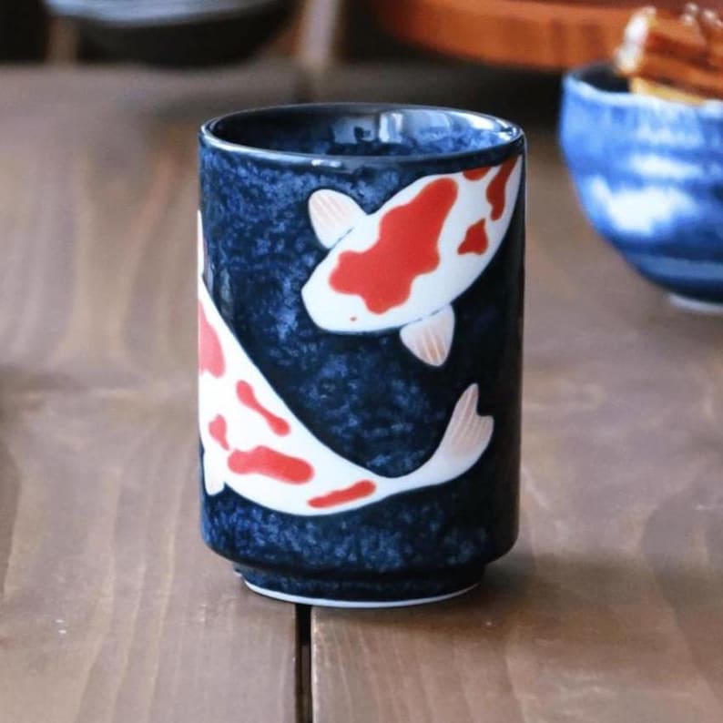 K&ouml;nnte beinhalten: Ein Keramikbecher mit dunkelblauem Hintergrund und einem Koi-Fisch-Design. Die Koi-Fische sind wei&szlig; mit roten Markierungen und pfirsichfarbenen Flossen. Der Becher ist zylindrisch und steht auf einer Holzoberfl&auml;che.