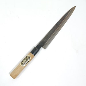 May include: A Japanese-style knife with a long, slender, dark blade. The handle is light-colored wood with a black band near the blade. A gold and black label is affixed to the handle. The knife is designed for precision cutting.