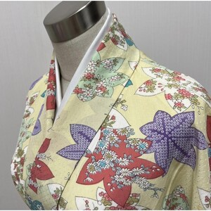May include: Close-up of a yellow kimono with a white collar. The kimono features a floral pattern in red, purple, and white. The fabric has a subtle sheen, and the design is intricate and detailed. The garment is displayed on a mannequin.