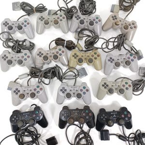 Pode incluir: Uma coleção de comandos com fios Sony PlayStation 2 em várias cores, incluindo cinza, bege e preto. Os comandos estão dispostos com os seus cabos e conectores visíveis. A imagem mostra uma variedade de acessórios de jogos retro.