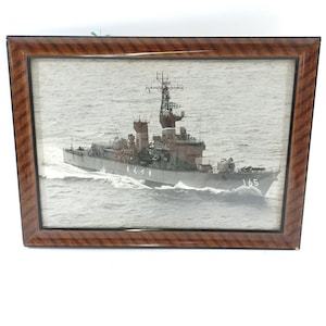 以下が含まれることがあります： 海上の軍艦の白黒写真が額装されています。船は水面を移動し、航跡を作っています。木製のフレームは、暗く模様のある仕上げです。船体番号は165です。