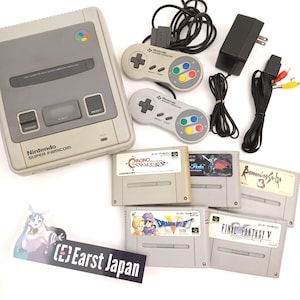 May include: A gray Nintendo Super Famicom console with two controllers, power adapter, and game cartridges. The cartridges include "Chrono Trigger", "Dragon Quest V", and "Final Fantasy V". The console and controllers are light gray.