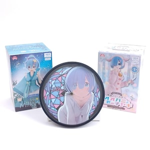 Puede incluir: Un reloj negro con un diseño de vitral colorido que presenta un personaje de anime con cabello azul. El reloj está rodeado por dos cajas con personajes de anime en ellas. Las cajas están etiquetadas como "Milky Way" y "Re:Zero - Starting Life in Another World".