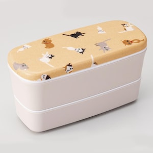 Op de afbeelding: Een roze bento-box met twee niveaus en een bruin deksel. Het deksel is versierd met een herhalend patroon van speelse kattenillustraties in verschillende poses en kleuren, waaronder zwart, bruin en wit. Ontworpen voor het meenemen van lunches.