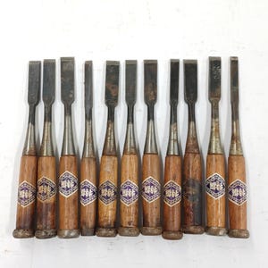 May include: A set of eleven wood chisels with metal blades and wooden handles. Each handle features a diamond-shaped purple and white label with Japanese text. The metal blades show signs of wear and oxidation, and the wooden handles have a natural finish.