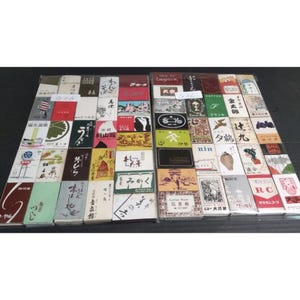 May include: A collection of rectangular matchboxes arranged in two grids, each featuring unique graphic designs, Japanese calligraphy, and various illustrations in diverse colors including red, green, black, and white, with some labels showing G06.