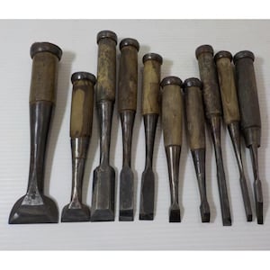 May include: A collection of nine wood chisels arranged in a row, each featuring a weathered wooden handle with a metal ferrule and a dark, oxidized steel blade of varying widths, ranging from wide flat edges to narrow, pointed tips.