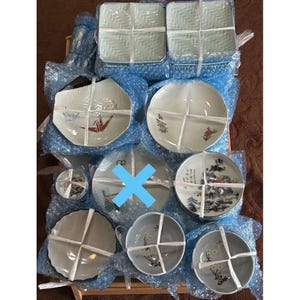 May include: A collection of various ceramic plates, bowls, and dishes, some square and some round, in shades of white, gray, and pale green. Some plates feature floral or landscape designs, while others have geometric patterns. The items are bundled together with white twine.