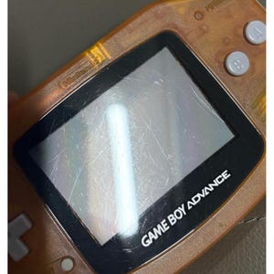 May include: An orange Game Boy Advance handheld console. The screen is scratched, and the text "GAME BOY ADVANCE" is visible. The Nintendo logo is also visible. The console has buttons on the right side and a directional pad on the left.