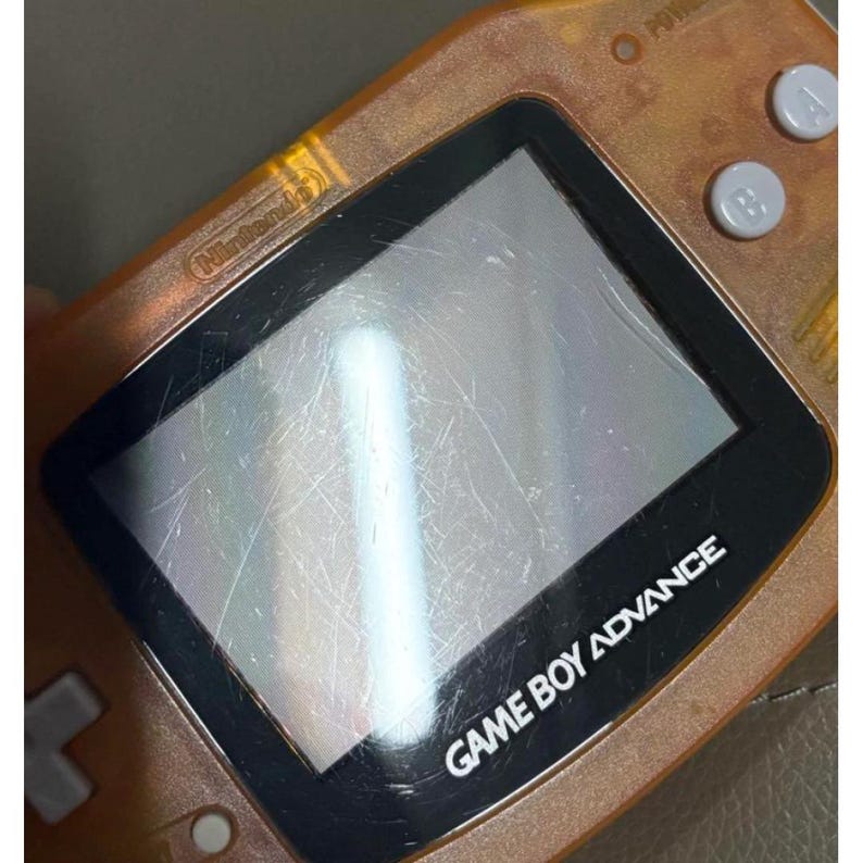 May include: An orange Game Boy Advance handheld console. The screen is scratched, and the text "GAME BOY ADVANCE" is visible. The Nintendo logo is also visible. The console has buttons on the right side and a directional pad on the left.