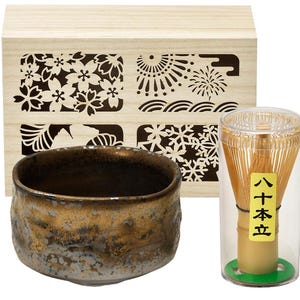 Op de afbeelding: Een Japanse theeceremonie set met een getextureerde, goud-bruine keramische kom, een bamboe klopper in een doorzichtige container en een houten doos met bloemmotieven. Voor het bereiden van matcha.