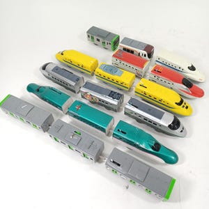 Puede incluir: Una colección de trenes de juguete en varios colores, incluyendo amarillo, verde, rojo y gris. Los trenes son de diferentes estilos, algunos con frentes puntiagudos y otros con diseños cuadrados. La imagen está tomada desde un ángulo alto, mostrando los trenes dispuestos sobre una superficie blanca.