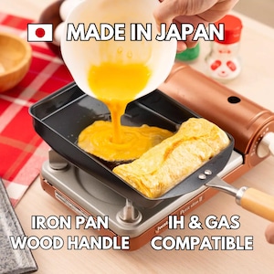 May include: A black iron pan with a wooden handle is shown on a silver and copper electric hob. The pan is filled with a yellow egg mixture and a rolled omelette. The hob has the text "Petit slim II" and "Iwat" printed on it. The text "MADE IN JAPAN", "IRON PAN", "WOOD HANDLE", "IH & GAS", and "COMPATIBLE" is also visible in the image.