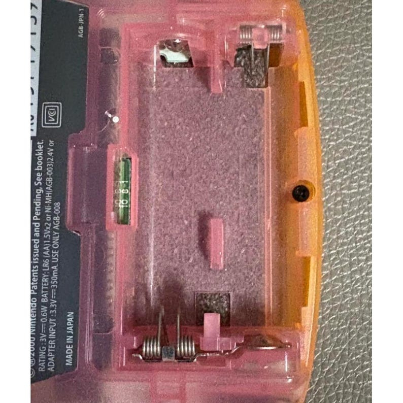 May include: Detailed view of the inside of a pink and orange handheld gaming device. The battery compartment is visible, with metal contacts and a green circuit board. Text includes "2000 Nintendo Patents issued and Pending" and "MADE IN JAPAN".