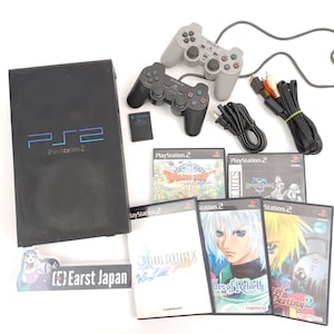 Puede incluir: Una consola PlayStation 2 negra con el logo "PS2" y el texto "PlayStation 2". Incluye dos mandos grises, varias carcasas de juegos, una tarjeta de memoria y cables. Las carcasas de los juegos presentan ilustraciones coloridas y títulos de juegos.