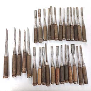 May include: A collection of thirty-one wood chisels with metal blades and wooden handles. The tools vary in width and length, featuring weathered steel blades and cylindrical wooden grips, arranged in rows on a plain white surface.