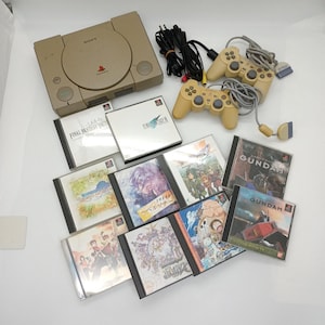Op de afbeelding: Een beige Sony PlayStation console met twee beige controllers en een verzameling PlayStation videogame-schijven, waaronder Final Fantasy VII, Final Fantasy VIII en Mobile Suit Gundam.