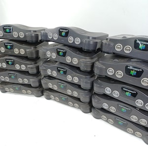 Puede incluir: Una pila de consolas de juegos Nintendo 64 grises. Cada consola tiene el logotipo de Nintendo 64 y puertos para mandos. Las consolas están apiladas, creando una exhibición de hardware de juegos retro.