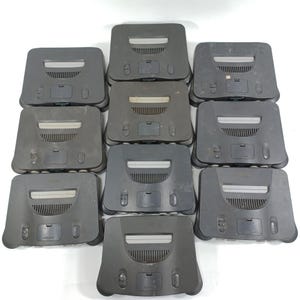 Puede incluir: Una colección de diez consolas de juegos Nintendo 64 grises. Cada consola tiene forma rectangular con una ranura central y varios puertos y botones. Las consolas están dispuestas en un patrón circular sobre un fondo blanco.
