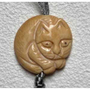 May include: A tan-colored, carved cat pendant with a detailed design. The cat is curled up, with its head and paws visible. The pendant is attached to a gray cord.