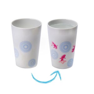 May include: Two white ceramic tumblers. One is empty, the other filled with water. Both feature a design of blue concentric circles and pink goldfish. The tumblers are cylindrical and have a smooth, glossy finish.
