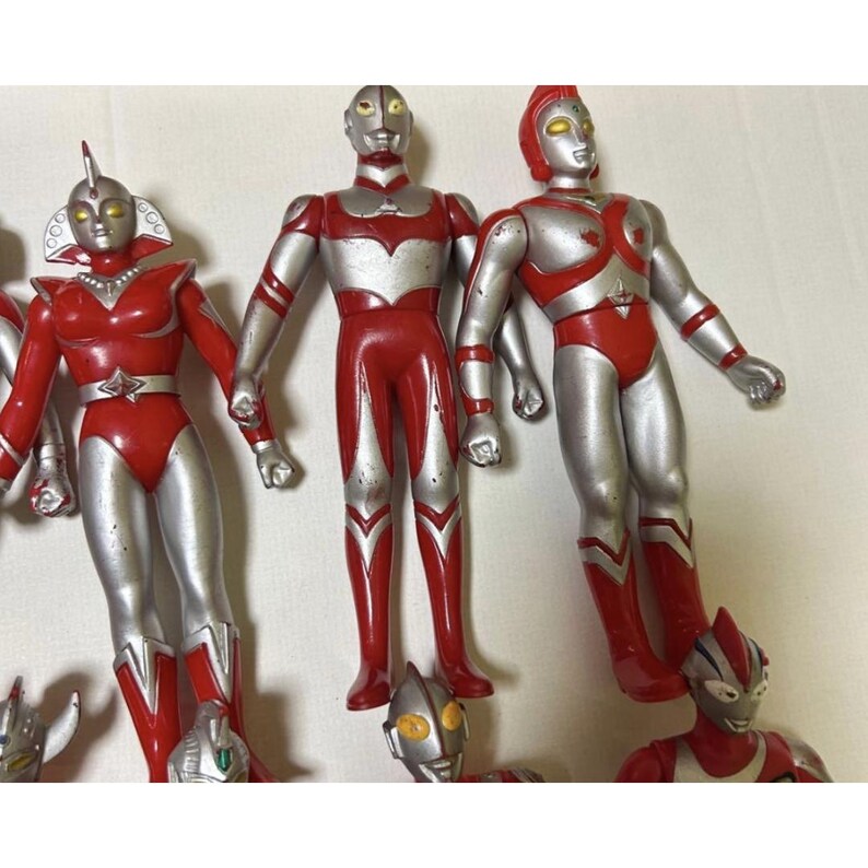 May include: A collection of red and silver plastic action figures featuring humanoid characters with stylized helmets and chest emblems. The figures are standing in various poses, displaying smooth surfaces and painted details on their bodies.