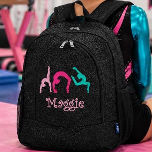 May include: A black glitter backpack with pink and teal gymnastics figures and the name "Maggie" in pink script. A pink water bottle with the text "EAT SLEEP GYMNASTICS REPEAT" and a black duffel bag with "GYMNAST" in pink are also visible.