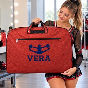 May include: A red glittery garment bag featuring a horizontal black zip pocket across the top. The centre of the bag displays a dark blue graphic of a cheerleader performing a jump, with the name VERA printed in dark blue block letters below.