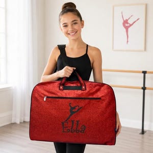 May include: A red glittery garment bag featuring a black zip, black handles, and a black shoulder strap. The front displays a black silhouette of a dancer in a leap pose above the name Ella written in a decorative black script font.