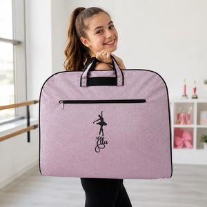 May include: A light purple garment bag with black trim and handles. The front features a black graphic of a ballet dancer in a pose above the name Ella written in a cursive script. The bag has a horizontal zippered pocket across the top.